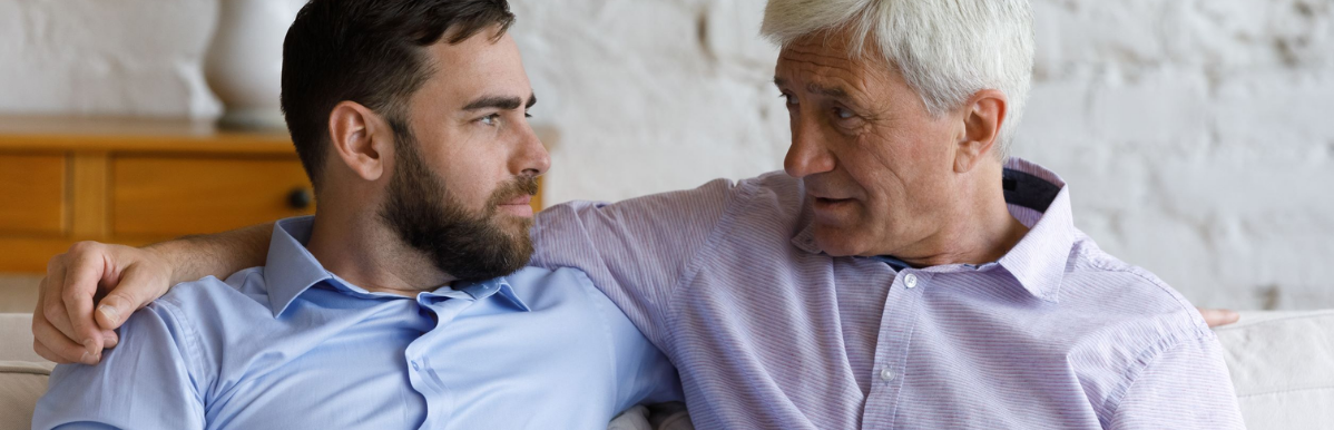 A younger man and an older man sit together on a couch, turned toward each other in close conversation. The older man, with gray hair, speaks while looking directly at the younger man, who listens with a serious, attentive expression. Above the photo, bold dark blue text on a light purple background reads: "When to Correct Your Loved One, and When to Let It Go."